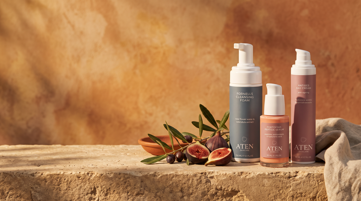 Three Aten skincare bottles arranged on a rustic stone surface with figs and olive branches against a warm textured backdrop. The set includes a tall blue bottle with a white pump, a smaller peach-toned bottle with a white pump, and a tall mauve-pink bottle with a white pump.